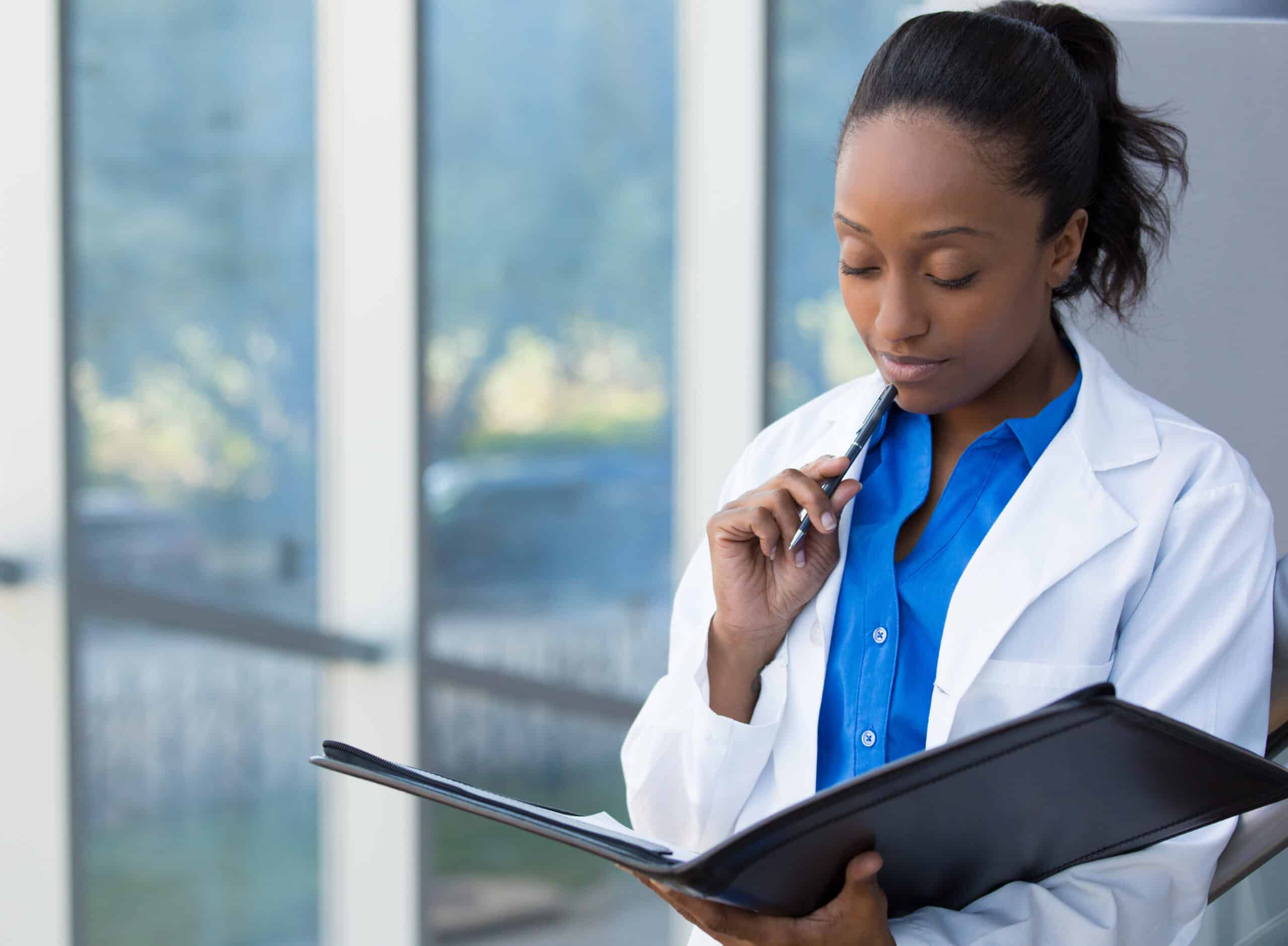 Closeup portrait of friendly, thinking confident female doctor, healthcare professional with labcoat, holding pen to face and holding notebook pad. Isolated hospital clinic background.